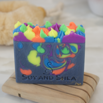A dark blue soap bar wiht neon coloured drops and piped top sits on a wood soap dish with a blurred background