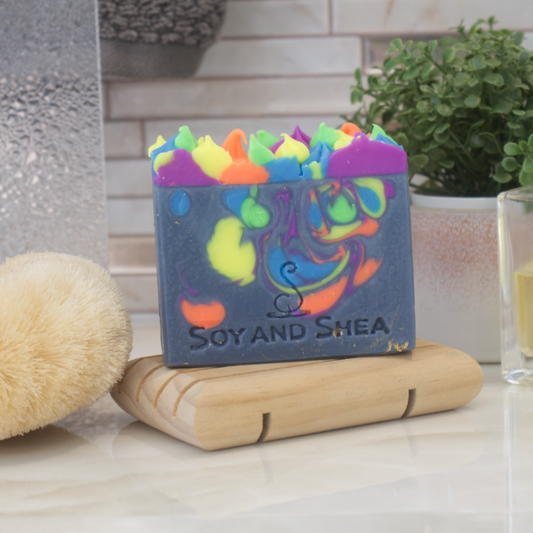 A Soap bar sits on a wooden soap dish on a bathroom counter top next to a body brush and potted plant.  The soap has a dark blue base with neon colour swirls.  These colours also appear as piped soap on the top of the bar