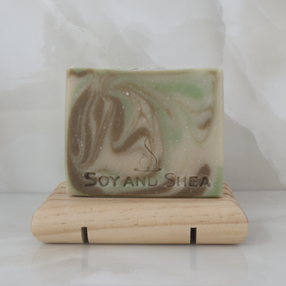 A soap with a light green colour and brown and bright green swirls stands upright on a wooden soap dish surrounded by marble tiles