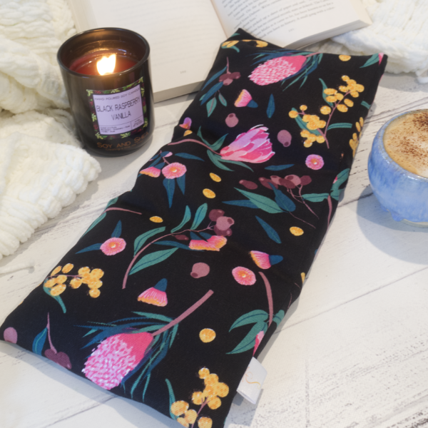 A long rectangular heat pack lays across a wooden surface with the corner resting on an open book propped up on a white blanket.  To the side is a burning candle and a cup of coffee suggesting a cosy scene. The Heat Pack is black with illustrations of Golden Wattle, Pink Gum flowers, gum nuts and pink banksia flowers.