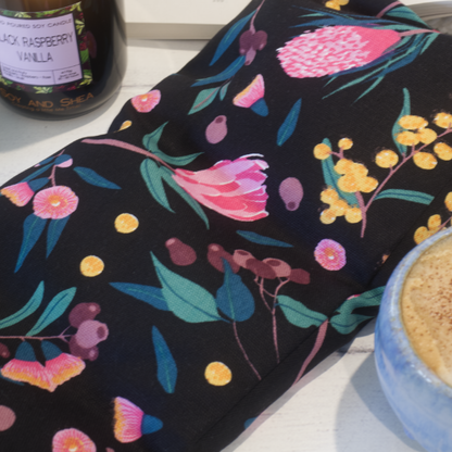 A close up of the Aussie Bush Heat Pack showing a fabric that is black with illustrations of Golden Wattle, Pink Gum flowers, gum nuts and pink banksia flowers.