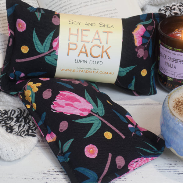 Two small rectangular heat packs are stacked upon each other and resting against a book. Beside them is a lit candle and cup of coffee. The heat packs have a black fabric with bright pink gum flowers and yellow wattle.