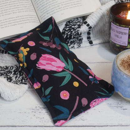 A small heat pack rests on a book  with a lit candle and cup of coffee to the right.  The heat pack is black with australian flowers including pink gum flowers, yellow wattle and gum nuts