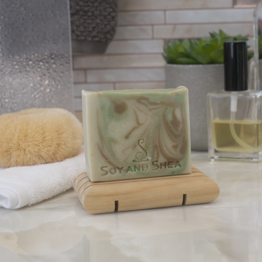 A bar of soap with a light green base and bright green and brown swirls sits on a wooden soap dish, with a white towel, a body brush to the side with a tiled background.