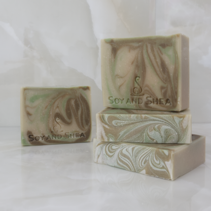 A collection of Aussie Bush Clay-tanical soaps sit on a marble tile background.  Two soaps lay flat and stacked on each other revealing a soap top wiht green and brown swirls.  A third soap stands upright on these showing a light earthy green soap wth brown and bright green swirls throughout.   A fouth soap stands in the background with the same colours but different swirl design.
