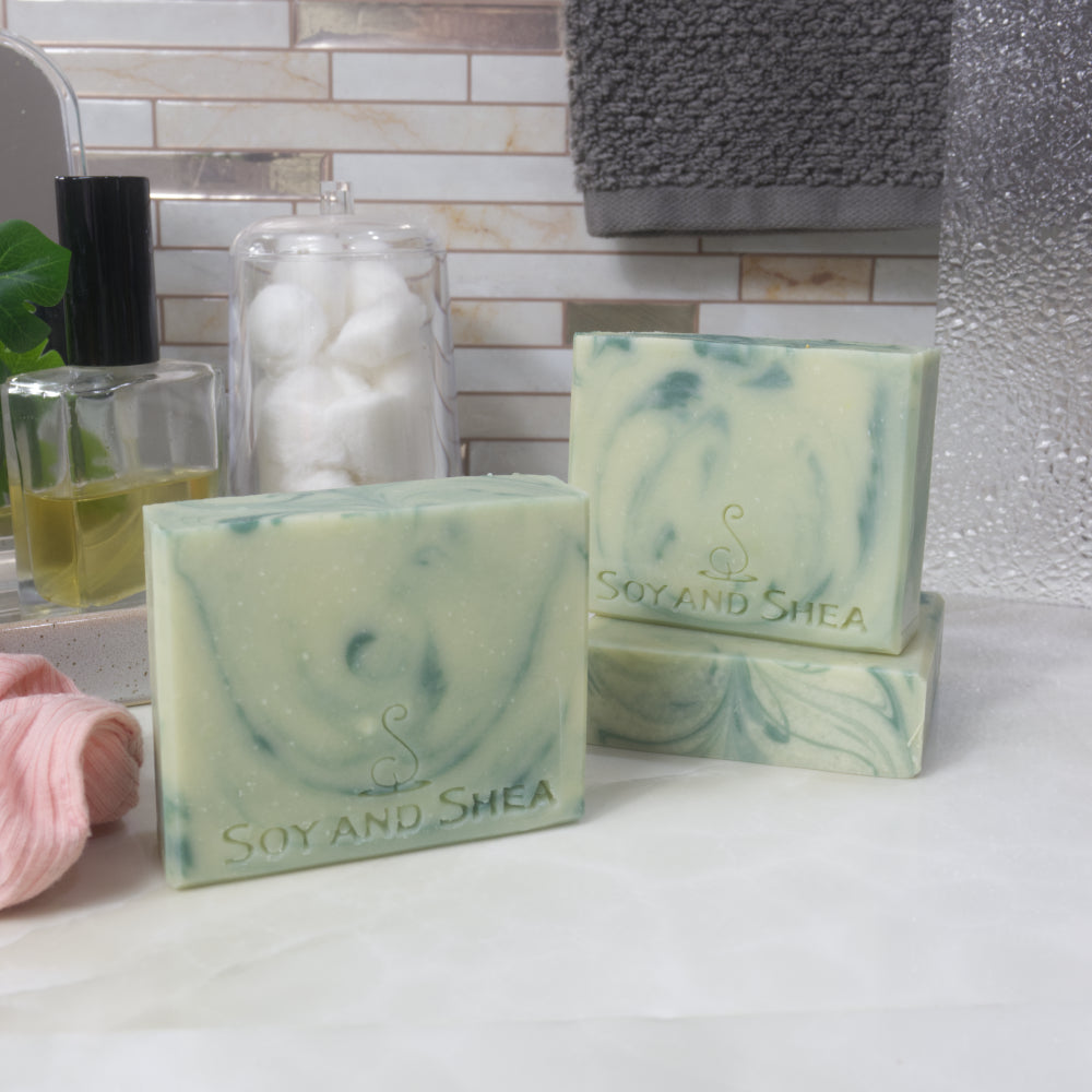 Three bars of soap are displayed on a batroom countertop.  To the front left, a soap bar stands upright, while to the back right another lays flat on the bench revealing a flat top with swirls of colour.  On this stands another soap.   All soaps are a pale green with varying streaks of dark green.
