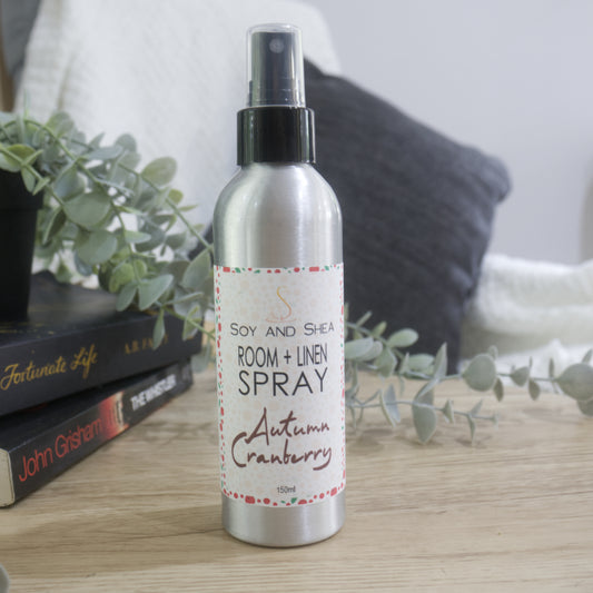 An aluminium bottle with black spray cap stands on a wooden surface next toa pile of books with a plant sitting on top of them.  A chair with white blanket and grey cushion is blurred in the background.   The bottle features a decorative product label identifying it as Room + Linen Spray, scented in Autumn Cranberry