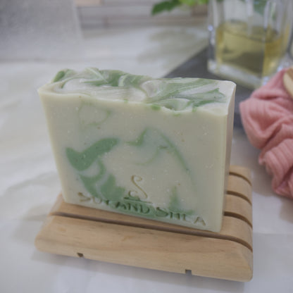 Pale Green Soap bar with dark green drops sits on a wooden soap rack with a blurred bathroom setting
