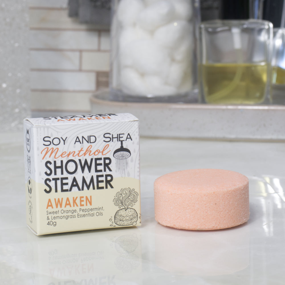 A boxed shower steamer and the steamer itself on a countertop in a bathroom setting. The box reads 'Soy and Shea Menthol Shower Steamer Awaken' and the round steamer is a pale peach-pink color.