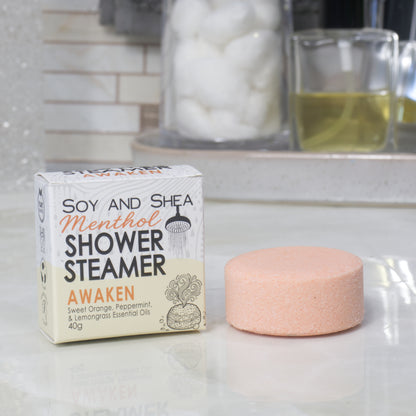 A boxed shower steamer and the steamer itself on a countertop in a bathroom setting. The box reads 'Soy and Shea Menthol Shower Steamer Awaken' and the round steamer is a pale peach-pink color.