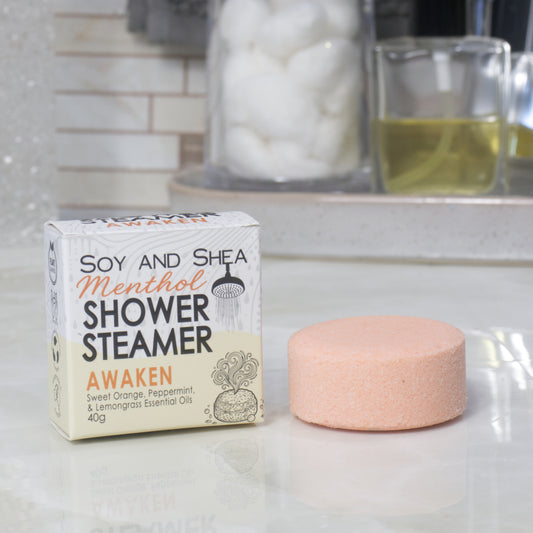 A boxed shower steamer and the steamer itself on a countertop in a bathroom setting. The box reads 'Soy and Shea Menthol Shower Steamer Awaken' and the round steamer is a pale peach-pink color.