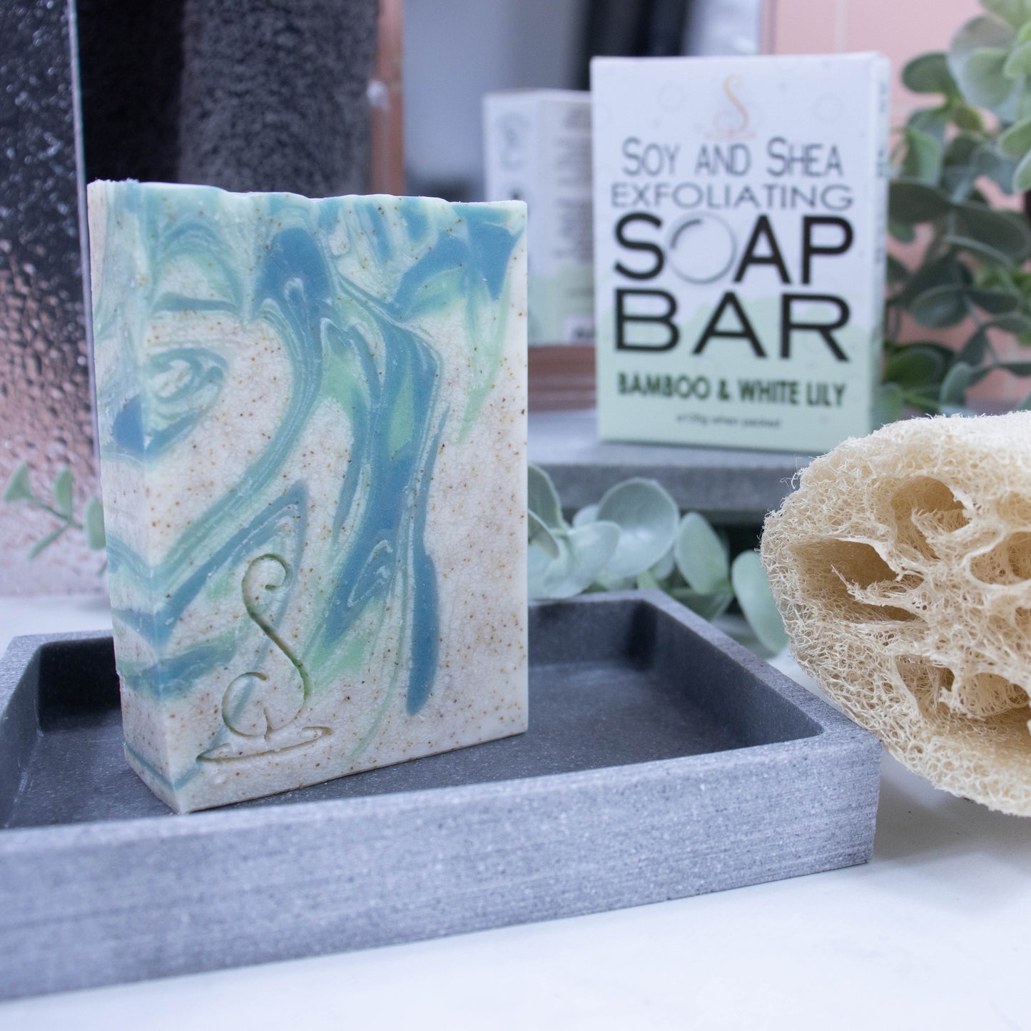 A tall rectangular bar of soap stands on a grey soap dish. The soap is a cream colour with light and dark swirls.  Throughout the soap are tiny brown speckles. Behind the soap is a shower screen and towel along with a box the soap was presented in. The box has a white top with pale green bubble border.