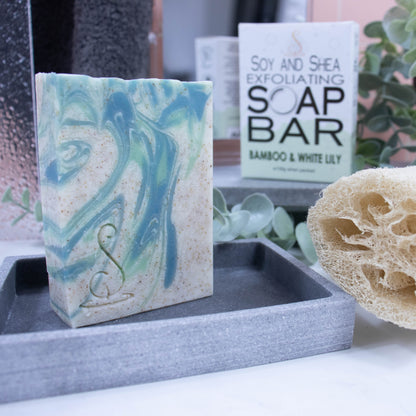 A tall rectangular bar of soap stands on a grey soap dish. The soap is a cream colour with light and dark swirls.  Throughout the soap are tiny brown speckles. Behind the soap is a shower screen and towel along with a box the soap was presented in. The box has a white top with pale green bubble border.