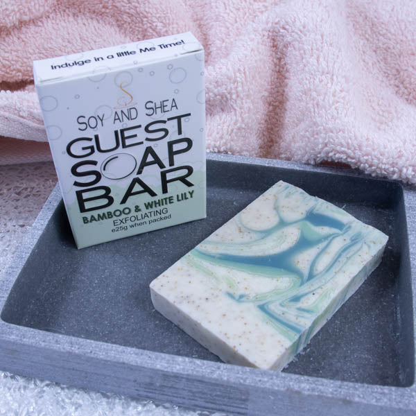 A grey stone soap dish sits next to a pink towel.  On the dish stands a white box with a border of ocean green bubble foam across the bottom and text GUEST SOAP BAR. Next to it lies a small piece of soap that is cream with light and dark green swirls with flecks of brown.