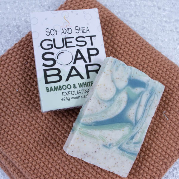 A rusty orange towel lies on a marble surface with a box labelled GUEST SOAP BAR.  Resting against the box is a small piece of soap that has a white base colour with light and dark green swirls along with flecks of brown.