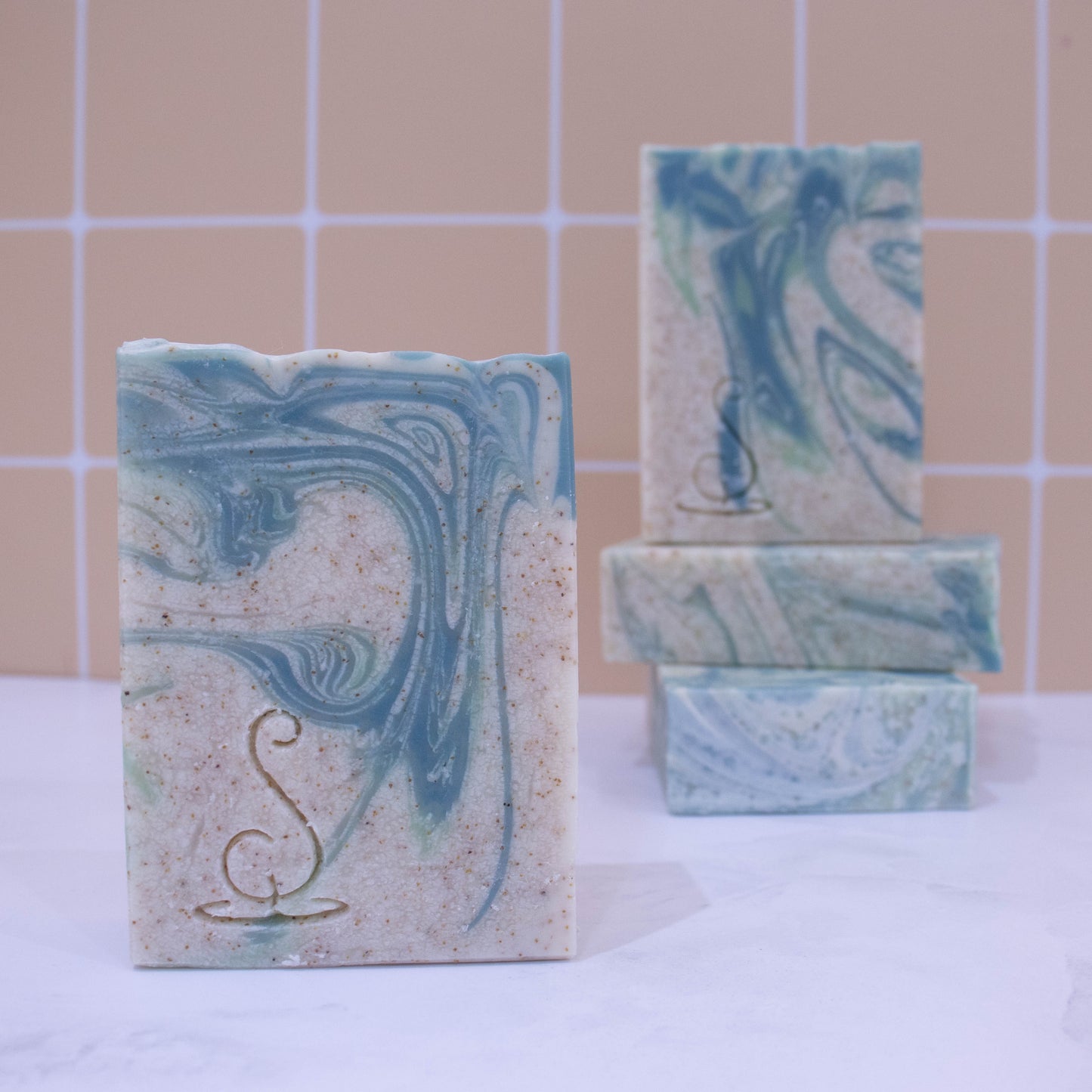 Four rectangular bars of soap sit on a marble bench each showing various design patterns from the same batch of soap. Two soaps lay flat but stacked upon each other showing a flat top.  A third soap stands on these while a fourth is presented in front of the stack. The soap is a cream colour with light and dark swirls.  Throughout the soap are tiny brown speckles. 