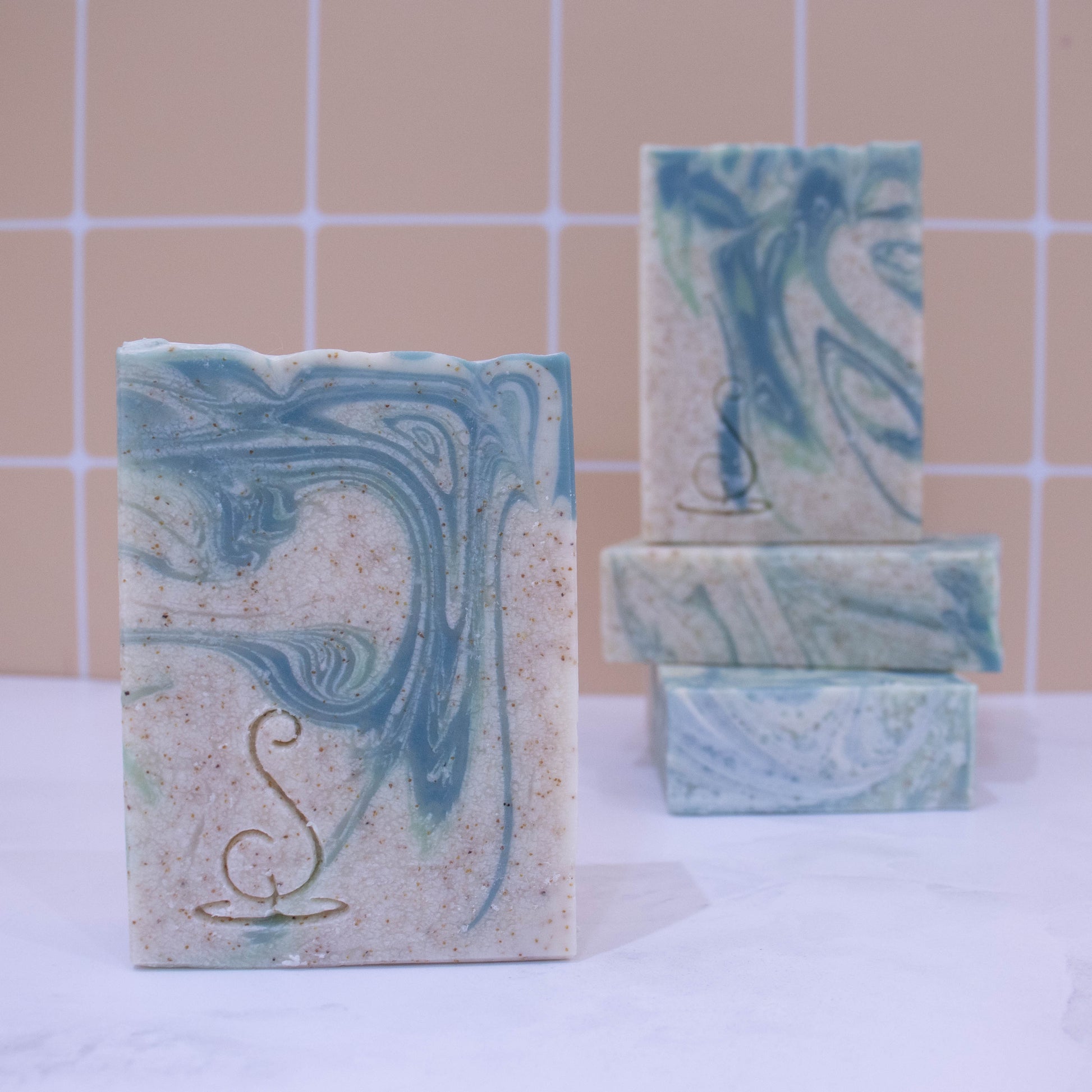 Four rectangular bars of soap sit on a marble bench each showing various design patterns from the same batch of soap. Two soaps lay flat but stacked upon each other showing a flat top.  A third soap stands on these while a fourth is presented in front of the stack. The soap is a cream colour with light and dark swirls.  Throughout the soap are tiny brown speckles. 