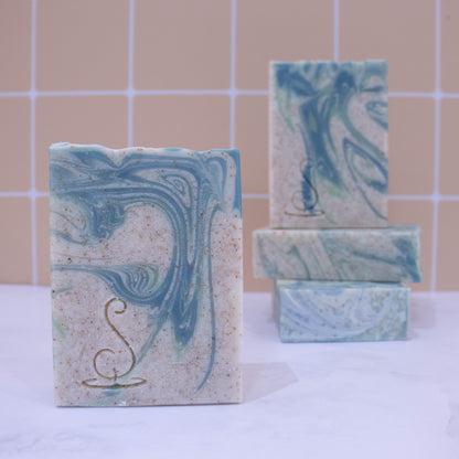 Four rectangular bars of soap sit on a marble bench each showing various design patterns from the same batch of soap. Two soaps lay flat but stacked upon each other showing a flat top.  A third soap stands on these while a fourth is presented in front of the stack. The soap is a cream colour with light and dark swirls.  Throughout the soap are tiny brown speckles. 