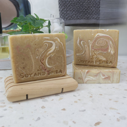 A selection of soap bars are displayed on a bathroom countertop.  One bar sits to the front right on a wooden soap dish, while another lays flat to the back right with another standing on top.  All soaps have a golden base colour with various swirls in bronze and white.  