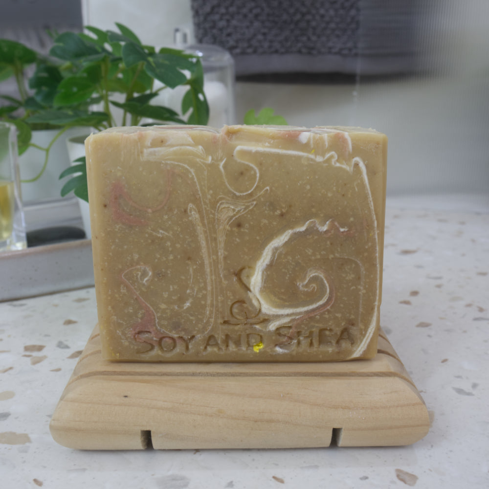 A bar of soap stands on a wooden soap rack next to a plant on bathroom counter top.  the soap has a golden base colour with swirls of white and bronze