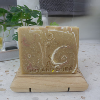 A bar of soap stands on a wooden soap rack next to a plant on bathroom counter top.  the soap has a golden base colour with swirls of white and bronze