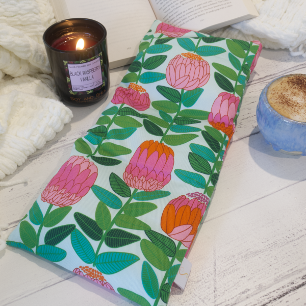 A long rectangular heat pack rests on a flat surface alongside a candle and a cup of coffee. Behind is a white blanket and open book.  The Heat Pack design features long green stems with leaves finished with a stylised banksia flower in shades of pink and orange all on a pale mint background.