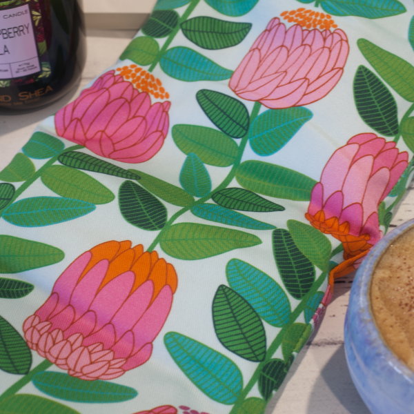 A close up image of the Banksia Heat Pack focusing on the fabric design featuring long green stems with leaves finished with a stylised banksia flower in shades of pink and orange all on a pale mint background.