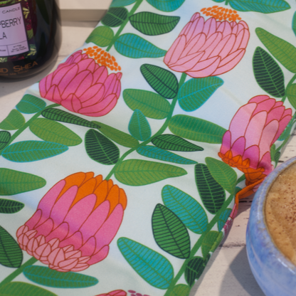 A close up image of the Banksia Heat Pack focusing on the fabric design featuring long green stems with leaves finished with a stylised banksia flower in shades of pink and orange all on a pale mint background.