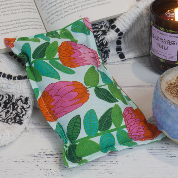 A Banksia Mini Heat Pack rests against a black and white blanket and open book. Beside it is a burning candle and filled coffee cup creating a cosy scene.  The Heat Pack has a mint background with dark green stems with leaves finishing with banksia flowers coloured in layers of bright pink and orange.