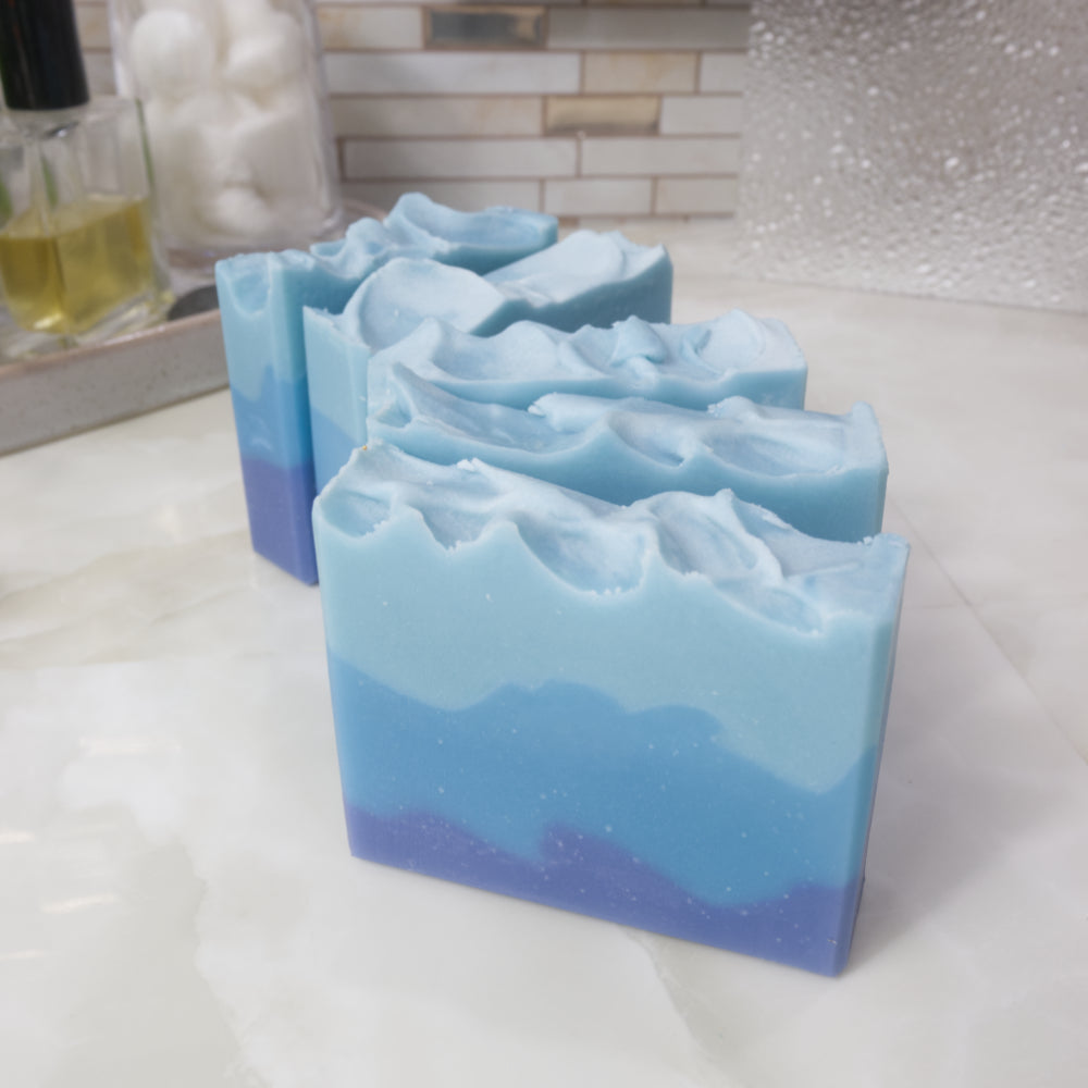 A selection of 5 blue soaps are presented on a bathroom counter.  They are lined up in front of each other so that their left edges touch and the right edge fans out in an arch.  The soaps feature three uneven layers going from dark blue on the bottom to light blue on top.