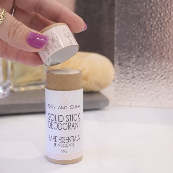A cardboard tube labelled SOLID STICK DEODORANT stands upright on a bathroom counter top with a blurred background.  A hand hovers above the tube holding the lid in the finger tips revealing  a cream balm inside