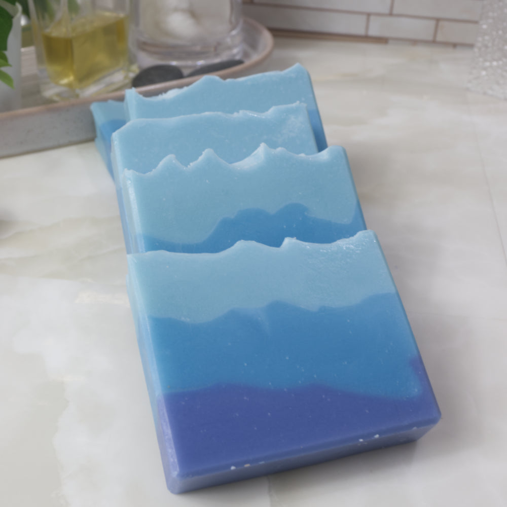 Five soap bars lay on a bathroom countertop in a domino effect.  The soaps feature three uneven layers going from dark blue on the bottom to light blue on top.