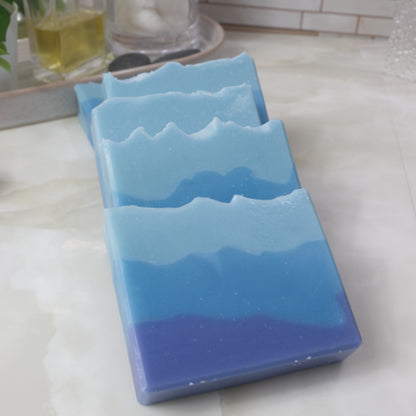 Five soap bars lay on a bathroom countertop in a domino effect.  The soaps feature three uneven layers going from dark blue on the bottom to light blue on top.