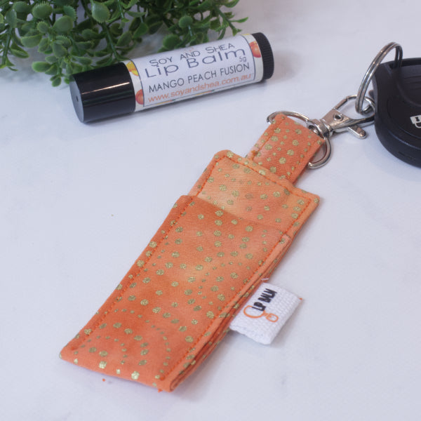 A lip balm holder lies on a wooden surface attached to a car key with a "lobster claw" clip.  To the side is a tube of lip balm surrounded by greenery.  The Clip Balm has a mottle orange background with sparkly gold dots.