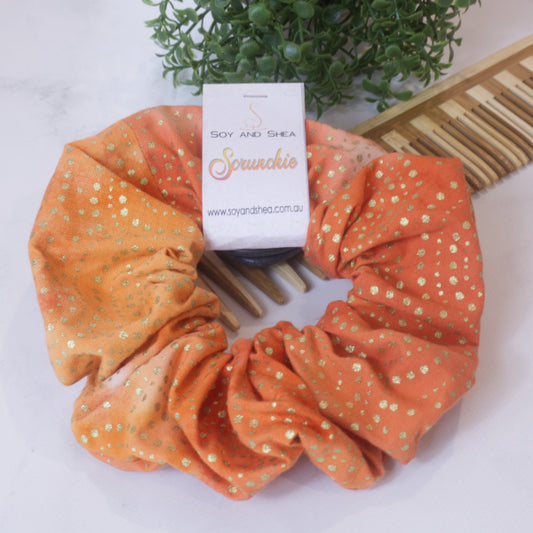 A colorful scrunchie sits on wooden surface resting against a wooden comb with greenery in the background.  A cardboard tag reads SOY AND SHEA SCRUNCHIE.    The scrunchie as a mottled orange background in the baticj style with shiny gold dots 