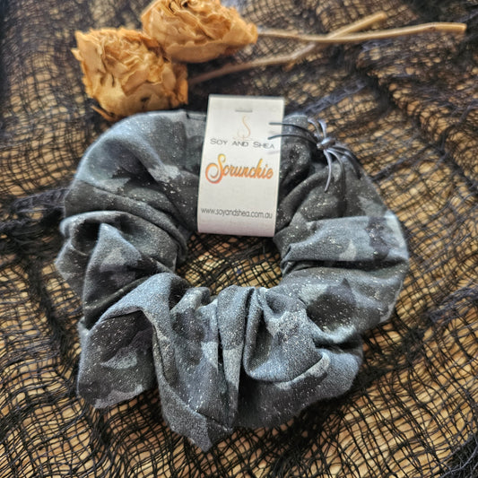 A grey scrunchie sits on wooden surface with a black mesh fabric covering it along side some dried rosesA cardboard tag reads SOY AND SHEA SCRUNCHIE.  The Scrunchie has a mottled black appearance with white flecks.  Solid white light and dark grey bat images cover the fabric.