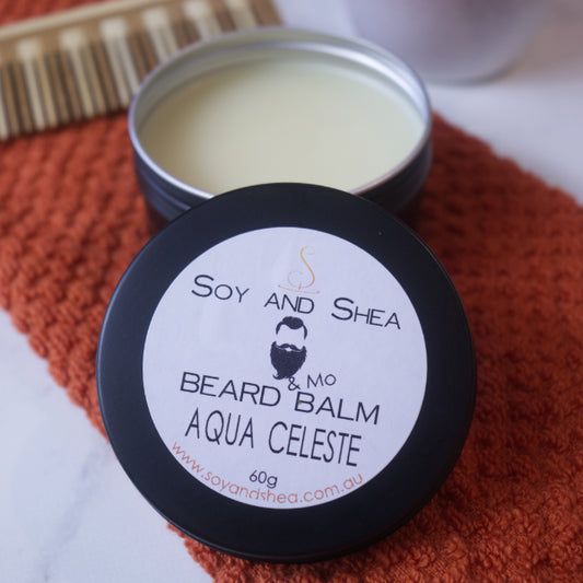 An open tin of Soy and Shea's Aqua Celeste Beard & Mo Balm displays its nourishing blend of natural ingredients. It sits on a textured orange cloth beside a wooden comb on a marble surface, perfect for conditioning beards.