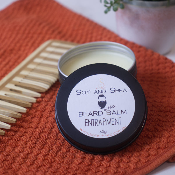 An open tin of Entrapment Beard & Mo Balm by Soy and Shea, crafted with natural ingredients, displays the moisturizing product inside. It sits on an orange knitted surface beside a wooden comb, with a small potted plant partially visible in the background.