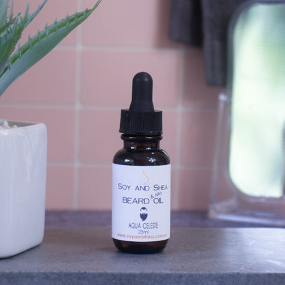 A small dropper bottle of "Aqua Celeste Beard & Mo Oil" from Soy and Shea rests on a gray countertop, promising to enhance your grooming routine. It sits beside a white ceramic planter with lush green foliage, against a backdrop of pink tiled wall and blurred mirror, highlighting this nourishing blend.