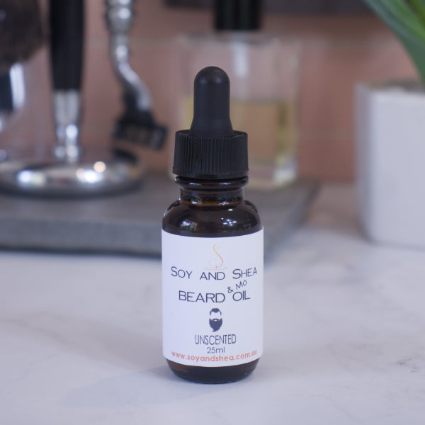 A small brown bottle labeled "Unscented Beard & Mo Oil" by Soy and Shea, featuring a dropper cap, rests on a marble surface. This nourishing 25ml blend is ideal for any grooming routine and is displayed with blurred bathroom items in the background.