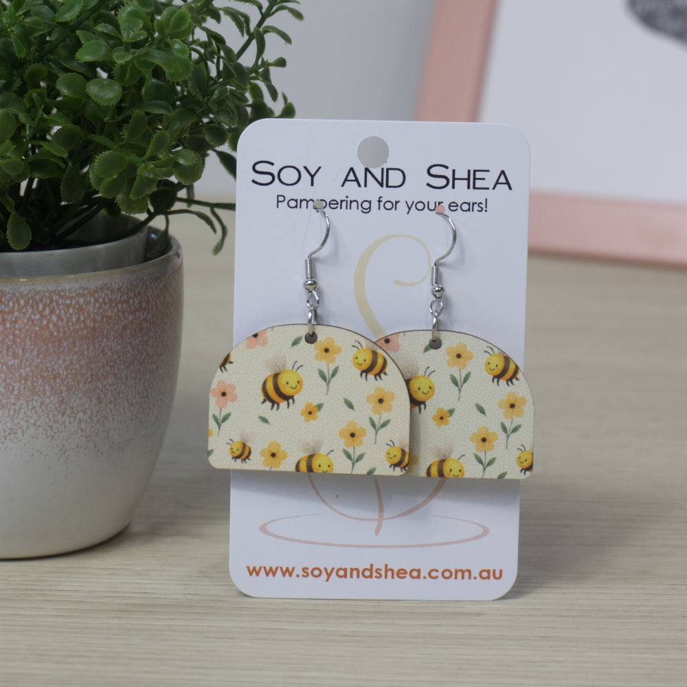 Arched shaped earrings with bee and flower pattern on a Soy and Shea card, with a plant in the background.