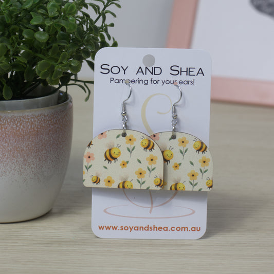 Arched shaped earrings with bee and flower pattern on a Soy and Shea card, with a plant in the background.