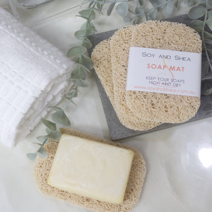 A flatlay image showing a collection of Beige Soap mats, displayed on a bathroom countertop along with a white towel, body brush and greenery.  Three soap mats are stacked upon each other in a fan shape on a grey stone tray with the top one featuring a product label.  A fourth mat lies to the bottom of the image with a cream soap lying on it.