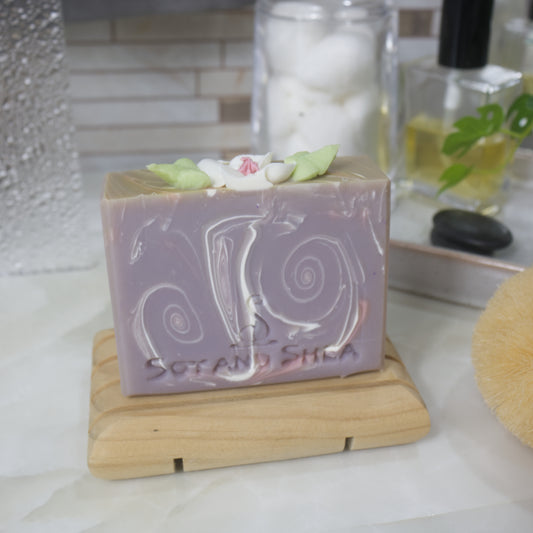 A bar of soap stands on a wooden soap dish on a bathroom countertop.  The soap has a light purple base colour with white and pink swirls throughout.  The top of the soap has a white flower with pink center and green leaves.  This has been made with soap
