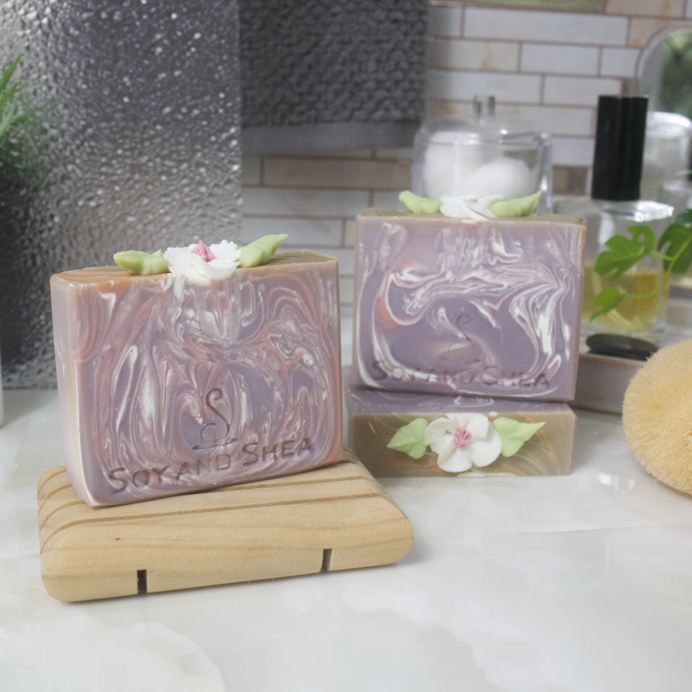 Three soaps are displayed on a bathroom countertop.  To the right, one soap lays flat showing the details of the soap top while another stands upon it.  The left front, is a third soap on a wooden soap rack.  The soaps have a light purple base colour with varying white and pink swirls throughout.  The top of each soap has a white flower with pink center and green leaves.  This has been made with soap