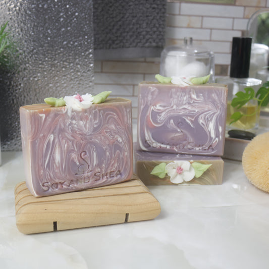 Three soaps are displayed on a bathroom countertop.  To the right, one soap lays flat showing the details of the soap top while another stands upon it.  The left front, is a third soap on a wooden soap rack.  The soaps have a light purple base colour with varying white and pink swirls throughout.  The top of each soap has a white flower with pink center and green leaves.  This has been made with soap