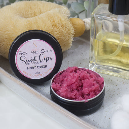 A round black tin sits on a stone tray along with a bottle of perfume and body brush.  The lid has been removed and is resting against the body brush revealing a cerise sugar scrub 