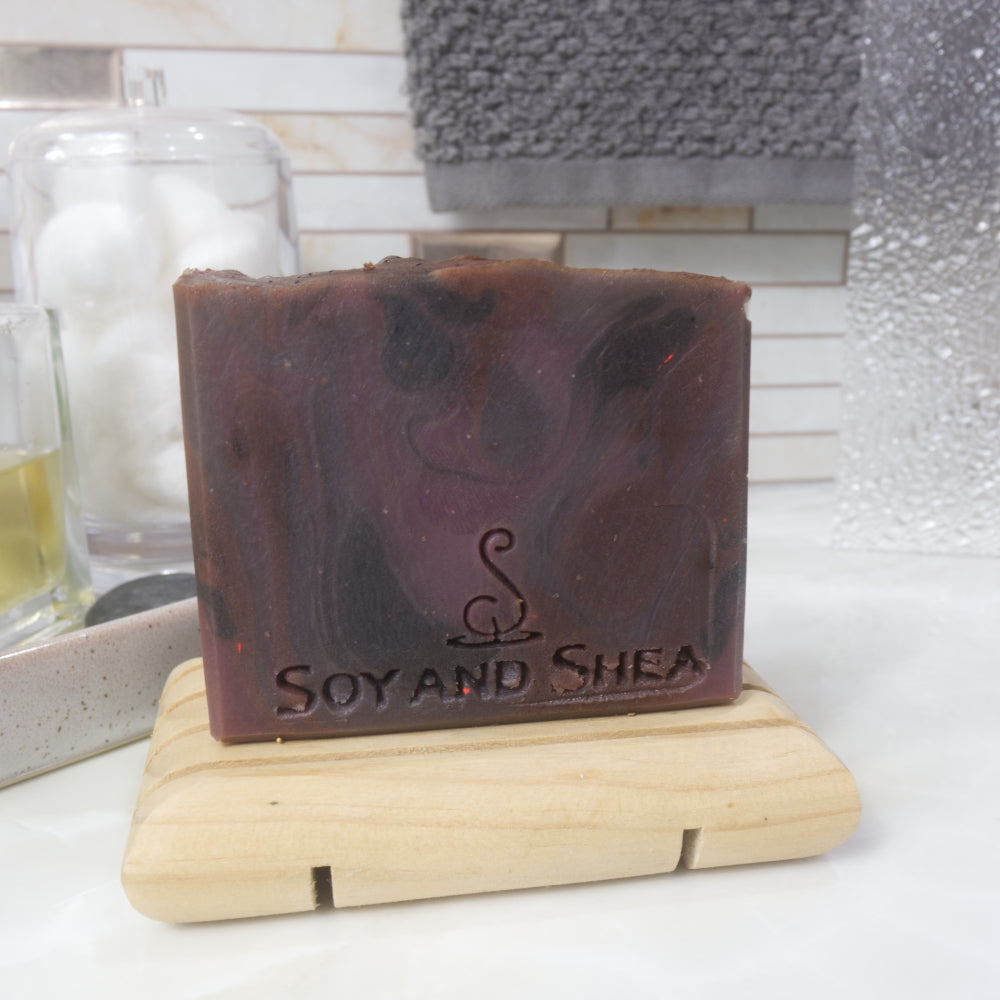 A rectangular bar of soap stands on a wooden soap rack on a bathroom counter top.  It has features a rich red, purple, black swirling pattern and a flat top
