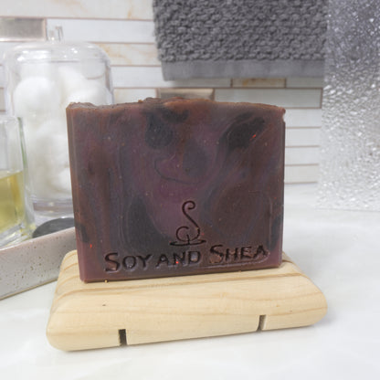 A rectangular bar of soap stands on a wooden soap rack on a bathroom counter top.  It has features a rich red, purple, black swirling pattern and a flat top