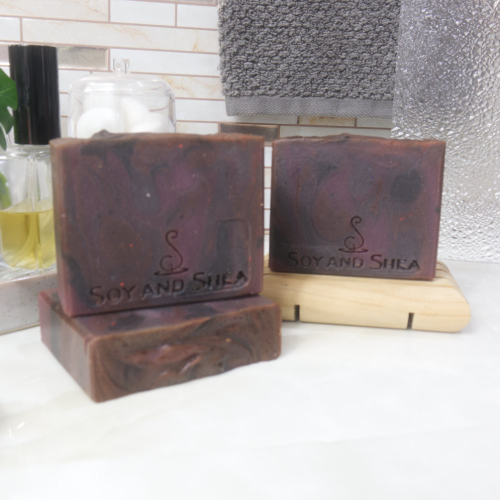 Three rectangular bars of soap are presented on a bathroom countertop next ot a bottle of perfume and a hanging towel.   One soap lies flat on the bench, with another standing upright on it.  To the back right, a third soap stands on a wooden soap rack.  THe soaps have a swirling pattern in dark shades of red and purples.